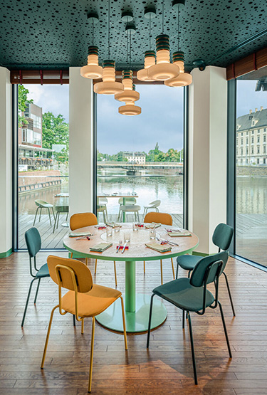 Przytulna restauracja z widokiem na rzekę, okrągłym stołem, żółtymi i zielonymi krzesłami oraz designerskim oświetleniem.